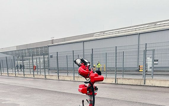 FireDos M1 fire monitor on a portable stand on an airfield apron, outside a hangar and behind a security fence.